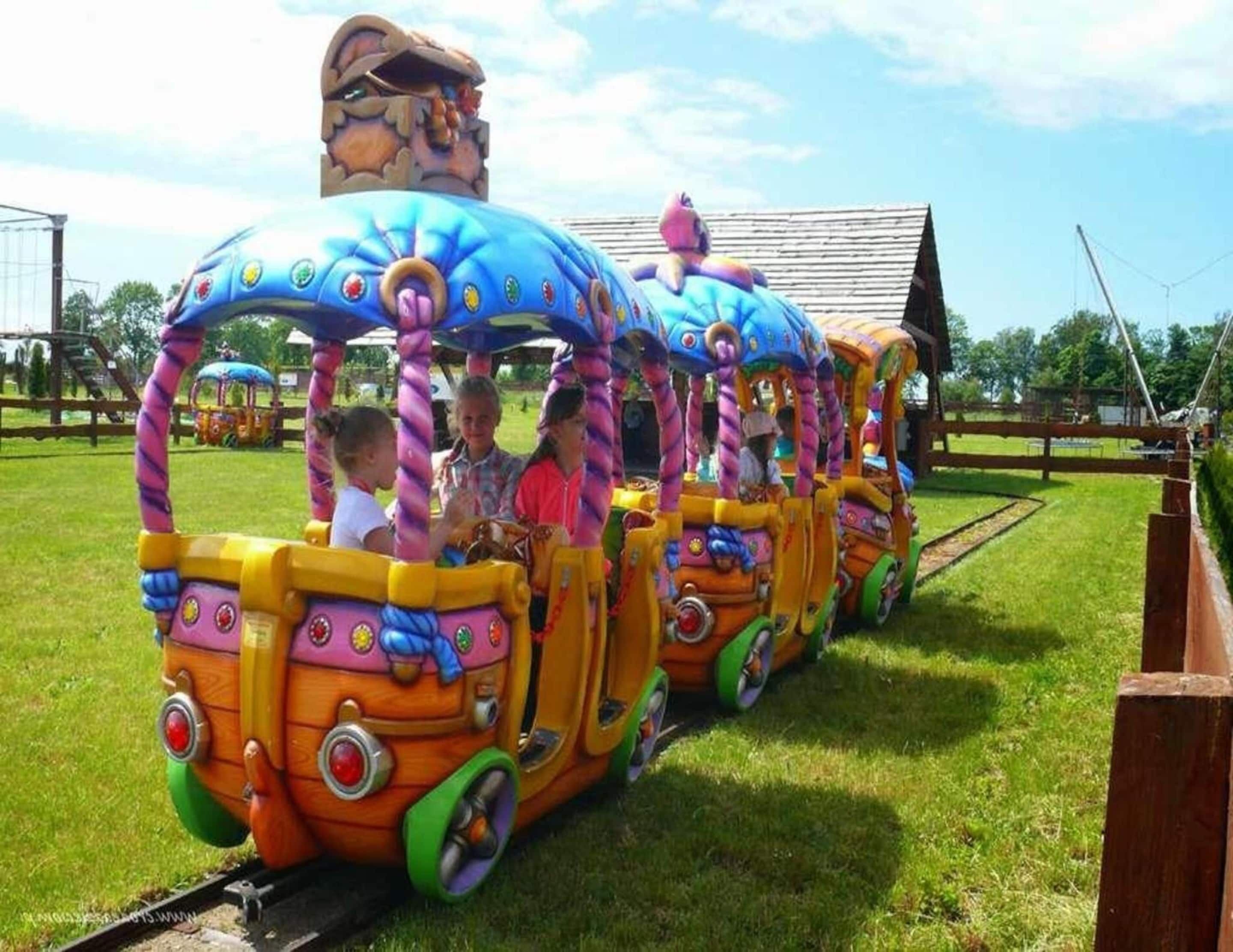 A colorful train ride with passengers, set on a grassy track with a wooden fence and a building in the background.