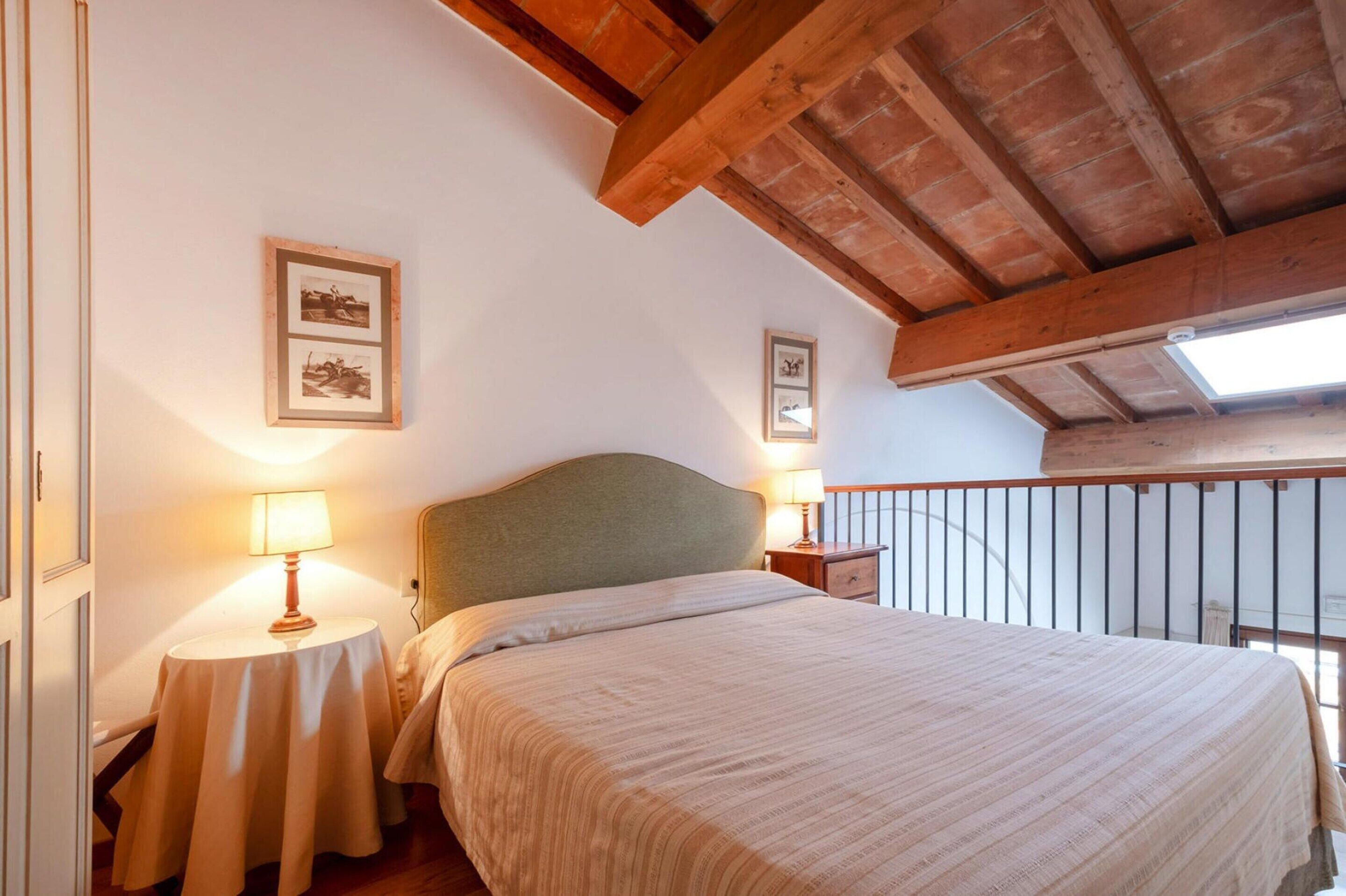 Un dormitorio con cama, dos mesitas de noche con lámparas, vigas de madera en el techo y cuadros enmarcados en la pared.
