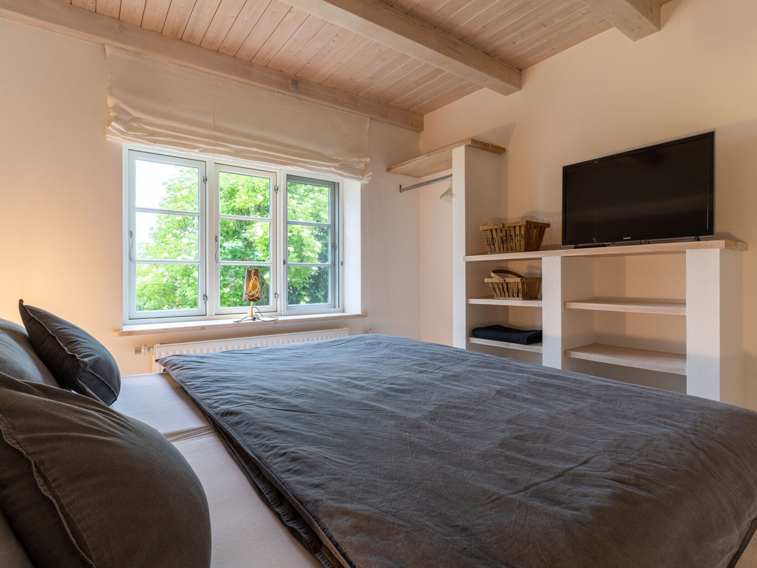 A bedroom with a large bed, a TV on a shelf, and a window with a view of greenery.