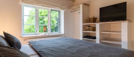 A bedroom with a large bed, a TV on a shelf, and a window with a view of greenery.