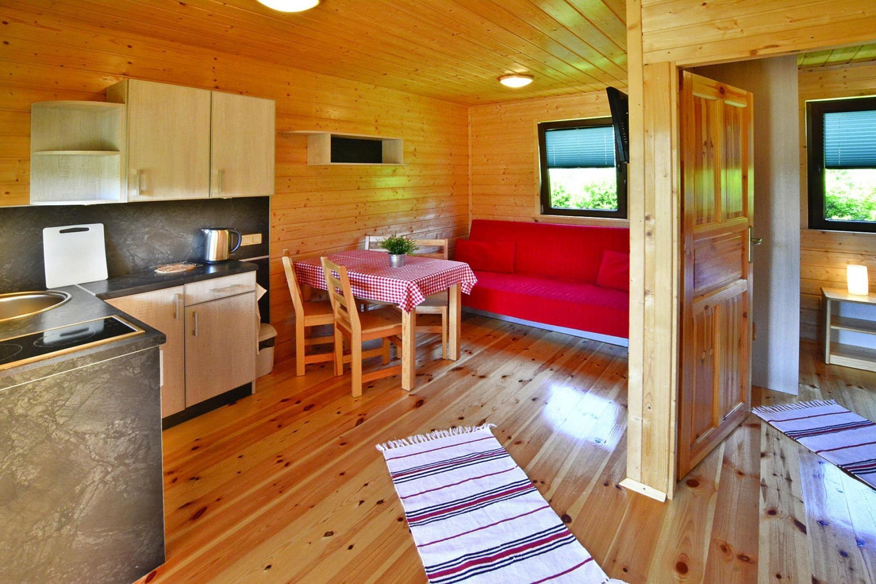 A wooden cabin interior with a kitchen, dining area, and a red sofa.