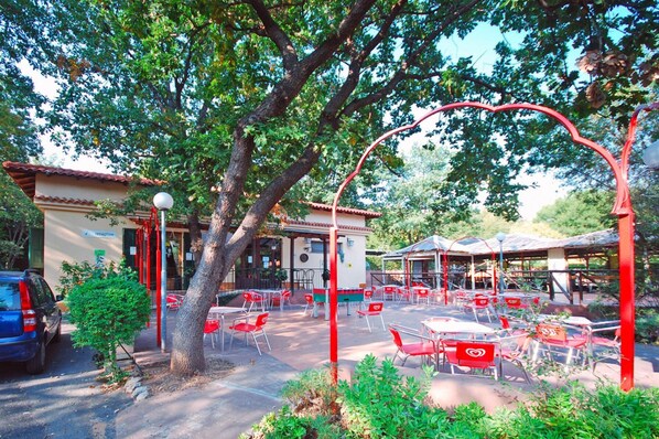 An outdoor cafe with red chairs and tables, surrounded by trees and a building with a reception area. - Holiday Village Caravelle Camping Vill, Ceriale (Ceriale)