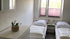 Ein Schlafzimmer mit zwei Betten, einem kleinen Fenster mit Jalousien, einem Nachttisch mit Lampe und einem Blumentopf auf der Kommode.