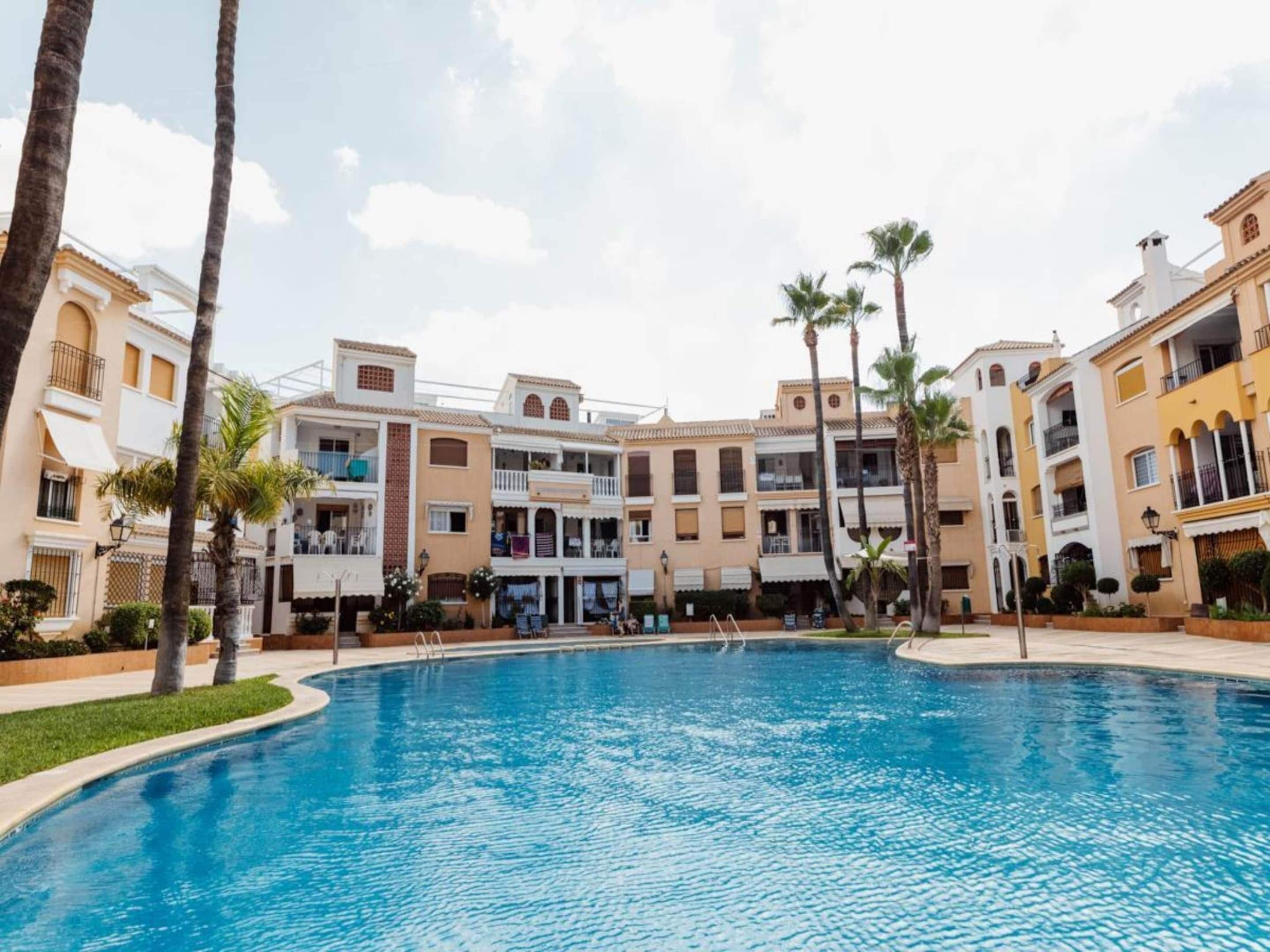 A swimming pool surrounded by multi-story buildings with balconies, palm trees, and a clear sky.