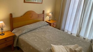 A hotel room with a wooden headboard, a bed with a floral-patterned bedspread, a nightstand with a lamp, and a window with curtains. - Apartment in Pueblo Salado, Mazarron (Mazarrón)