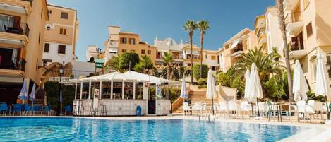 A swimming pool with lounge chairs and umbrellas, surrounded by multi-story buildings with balconies and palm trees.