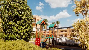 A playground with a swing set and climbing structures, surrounded by well-maintained hedges and palm trees, with buildings in the background. - Apartment in Pueblo Salado, Mazarron (Mazarrón)