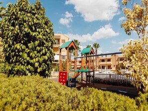 A playground with a swing set and climbing structures, surrounded by well-maintained hedges and palm trees, with buildings in the background. - Apartment in Pueblo Salado, Mazarron (Mazarrón)