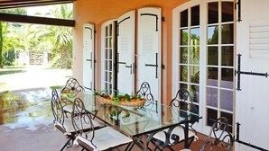 Une table à manger avec un plateau en verre, six chaises, un panier de fruits et une vue sur un jardin par de grandes portes-fenêtres.