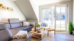 A modern living room with a sofa, coffee table, and chairs. There is a window with a view of buildings outside. - Apartment, Zingst (Zingst)