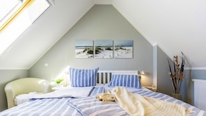 A bedroom with a sloped ceiling, a bed with striped bedding, a bedside table with a lamp, and a wall-mounted artwork depicting a beach scene. - Apartment, Zingst (Zingst)
