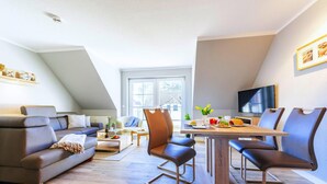 A modern living room with a sofa, a dining table, and chairs. There is a TV on the wall and a window providing natural light. - Apartment, Zingst (Zingst)