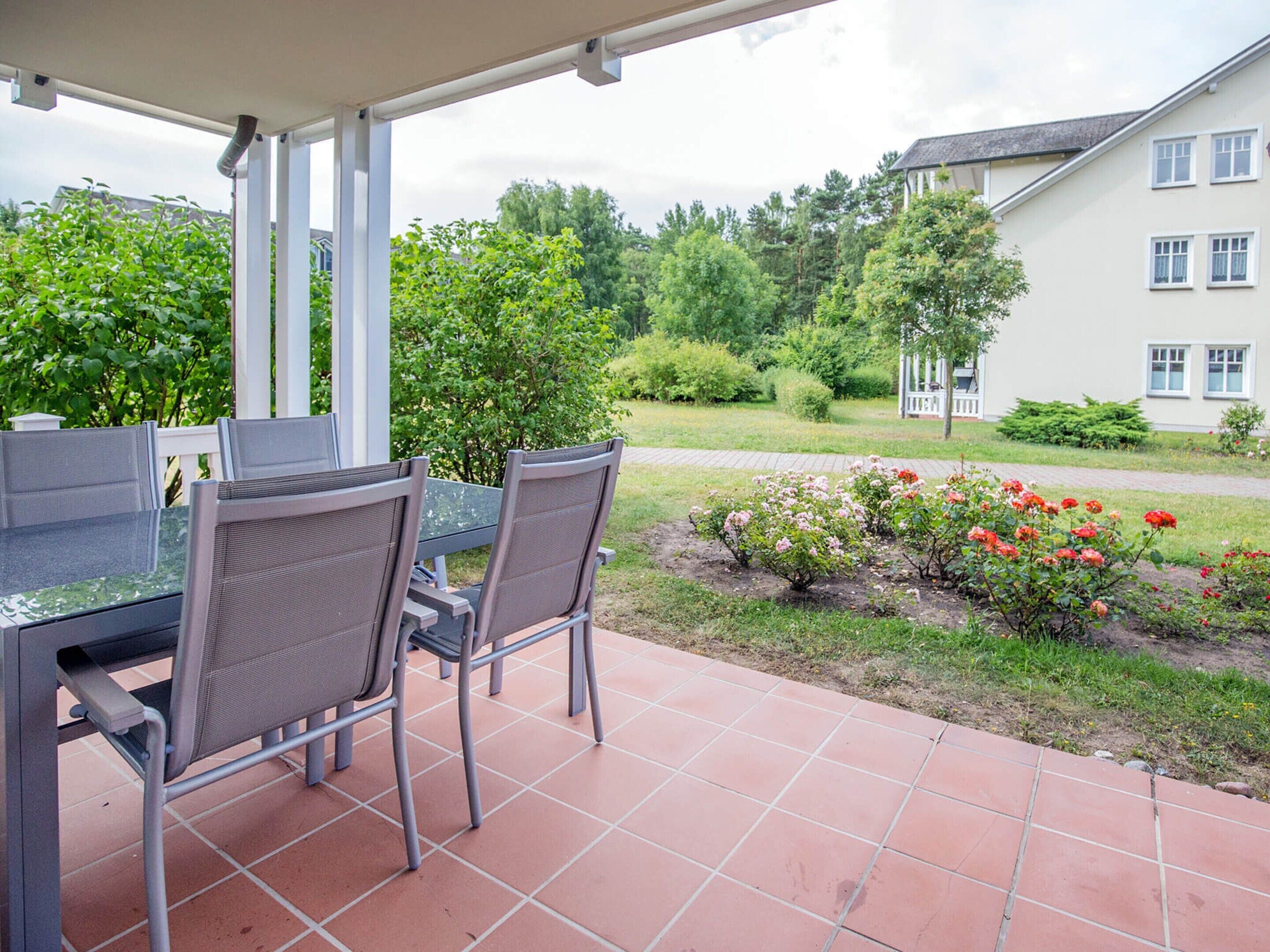 Um pátio externo com mesa e cadeiras, um jardim com rosas e um edifício ao fundo.