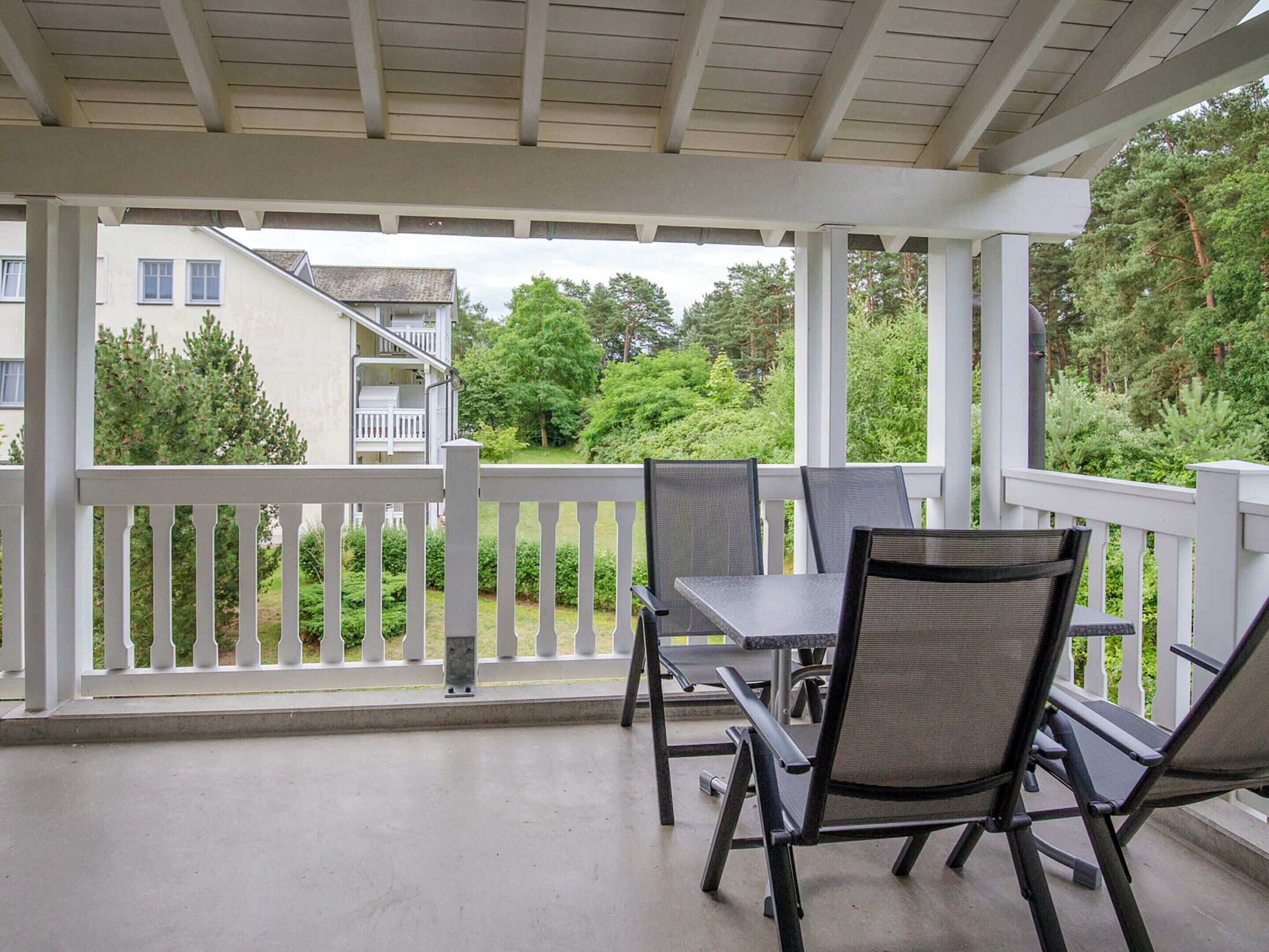 A covered patio with a table and chairs, white railings, and a view of a residential building and greenery.