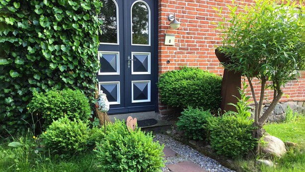 Una casa de ladrillo con una puerta azul oscuro, un pequeño jardín con arbustos variados y un buzón con el número 8.