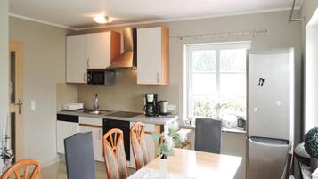 A modern kitchen with white cabinets, a microwave, a coffee maker, and a refrigerator. There is a dining table with chairs and a window providing natural light.