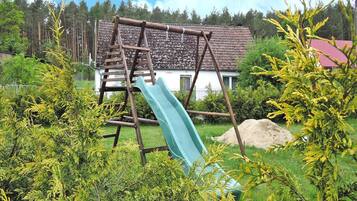 A wooden swing set with a slide in a grassy area with trees and a house in the background.