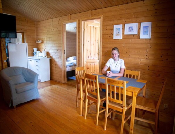 A cozy cabin with wooden walls and floors, a dining area with wooden chairs and a table, a kitchenette with a refrigerator and microwave, and a bed in the adjacent room. - Bungalow in Miedzyzdroje Near Baltic Beach (Międzyzdroje)