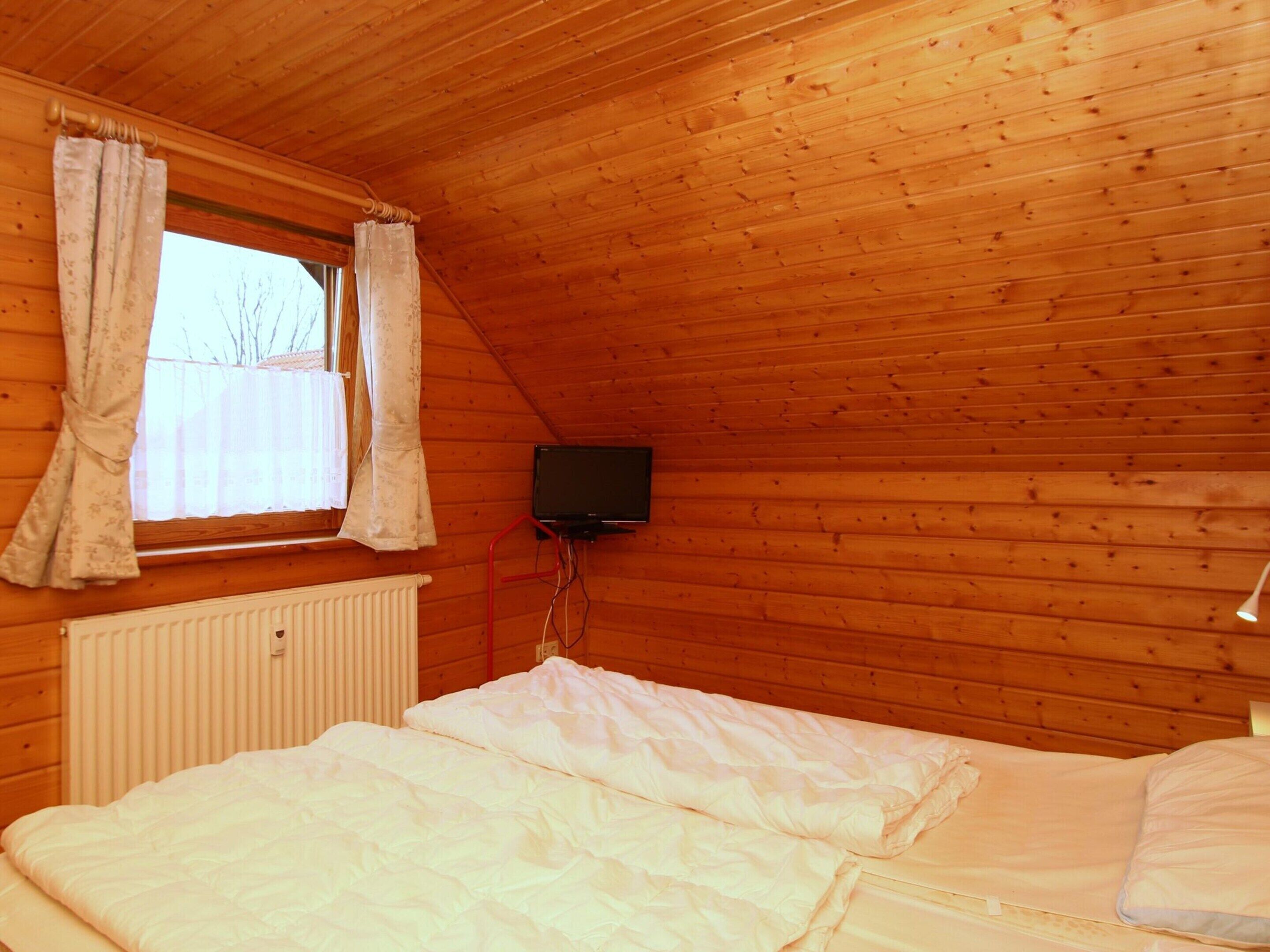 Ett sovrum med ett träpanelat tak, en säng med vita sängkläder, ett fönster med gardiner och en tv som är monterad på väggen.