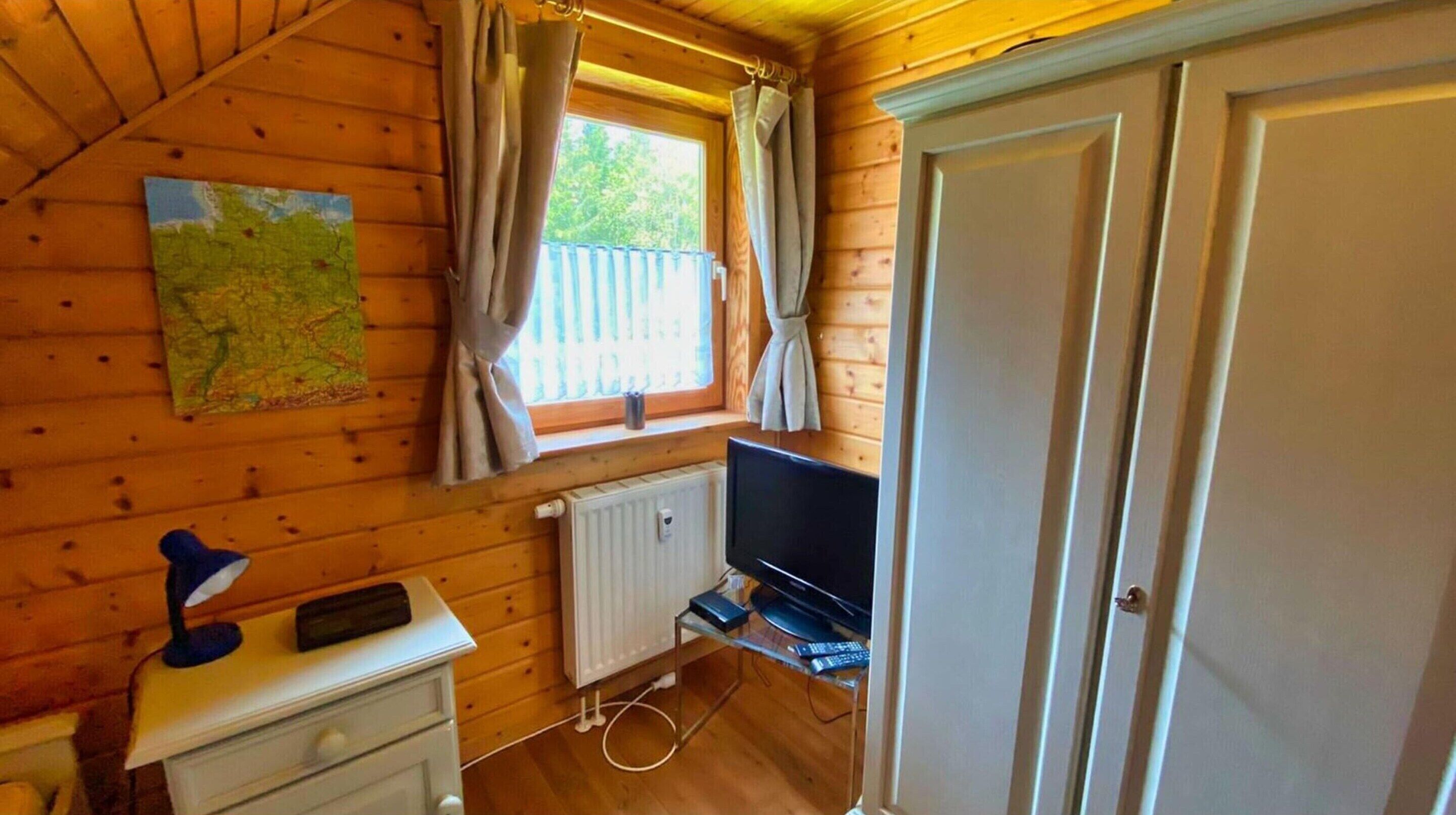 A wooden cabin room with a map on the wall, a window with curtains, a radiator, a TV, and a door.