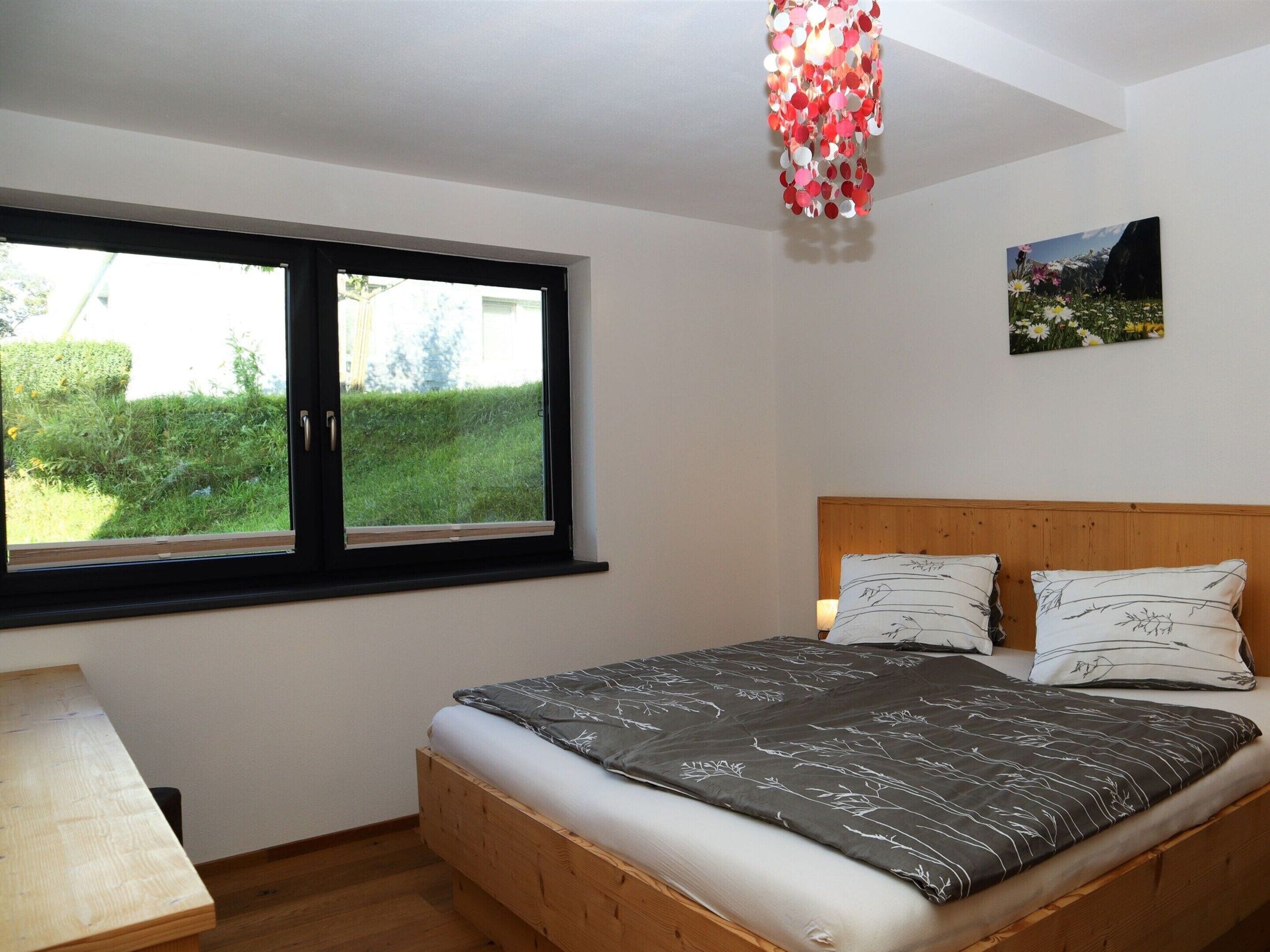 A bedroom with a wooden bed, a window with a view of greenery, and a wall-mounted picture.