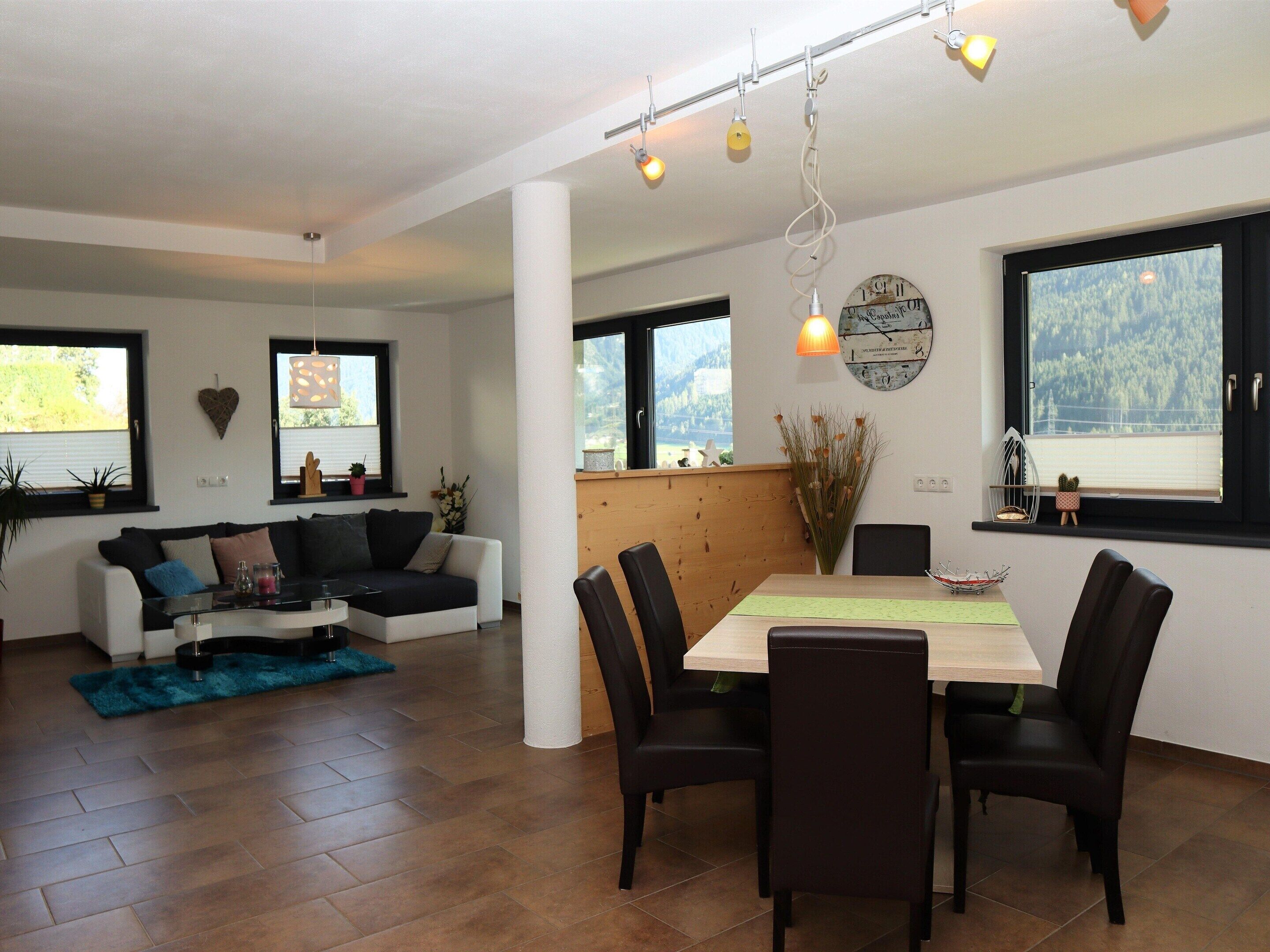 A modern living room with a sofa, dining table, and chairs. There are windows with views of greenery and a clock on the wall.