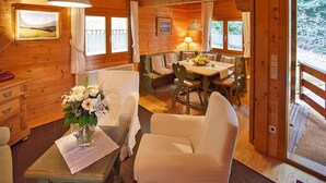 L’intérieur d’un chalet en bois comprend un coin repas, un canapé, une table ornée d’un vase de fleurs et une vue sur l’extérieur, visible à travers une porte.