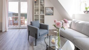 A modern living room with a grey sofa, a grey armchair, and a glass coffee table. There is a bookshelf with glass doors, a dining area with a table and chairs, and a balcony with a view. - Liebevoll Eingerichtete Ferienwohnung in Husum (Husum)