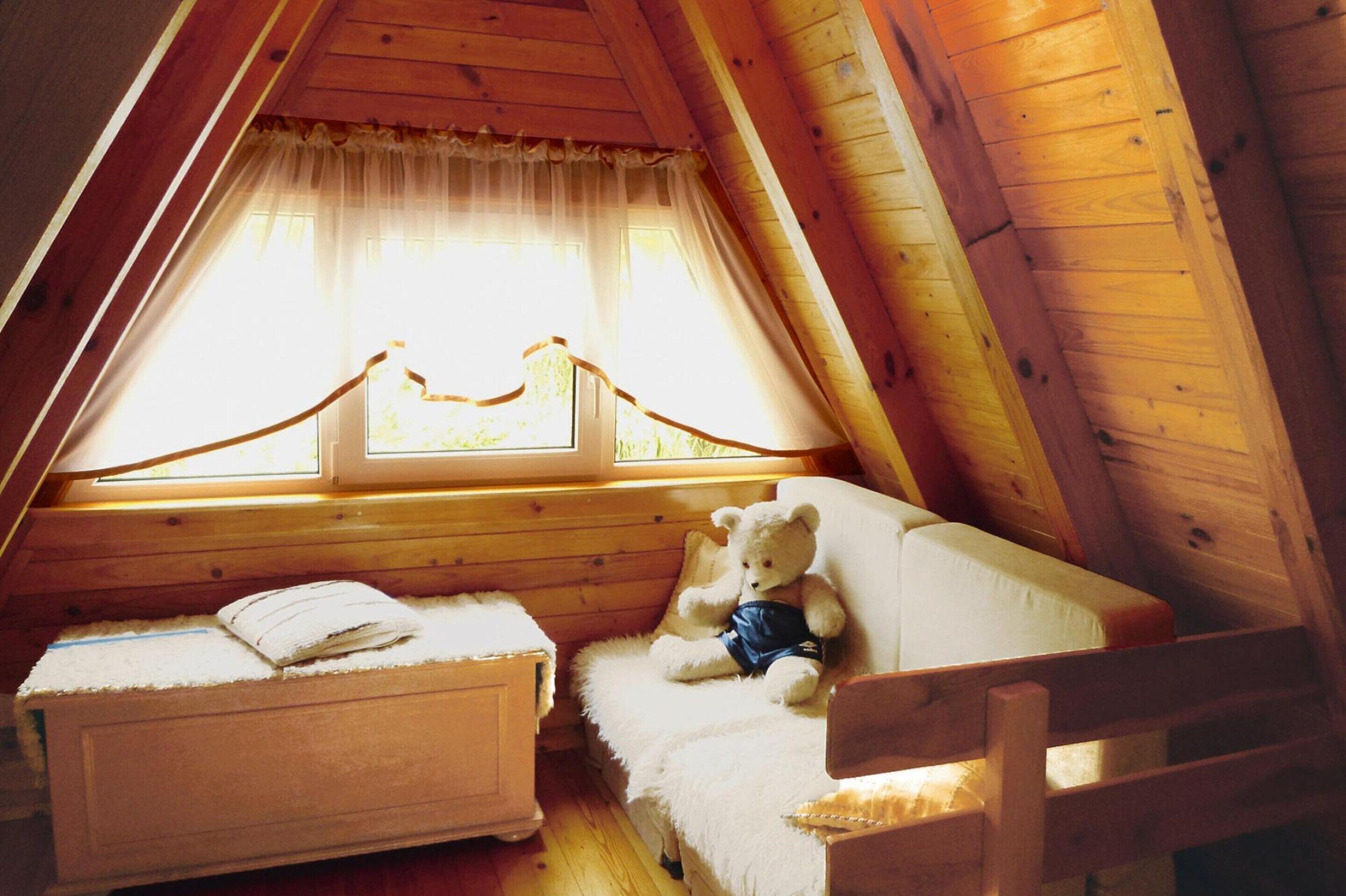 A cozy attic bedroom with a wooden sloped ceiling, a single bed with a white comforter, a small bedside table, and a teddy bear on the bed.