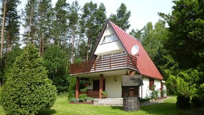 A small, single-story house with a red roof and white walls, surrounded by a well-maintained lawn and trees. - Holiday Home in Kashubia Near Lake (Suleczyno)