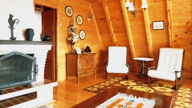 A cozy log cabin interior with a fireplace, two white armchairs, a small table, and wooden walls with decorative plates. - Holiday Home in Kashubia Near Lake (Suleczyno)