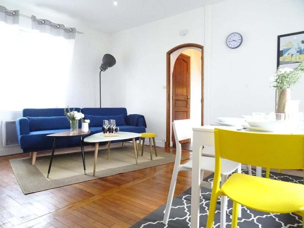 Uma sala de estar com sofá azul, mesa de centro, poltrona amarela, mesa de jantar com cadeiras, um relógio e uma porta.