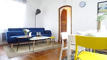 Uma sala de estar com sofá azul, mesa de centro, poltrona amarela, mesa de jantar com cadeiras, um relógio e uma porta.