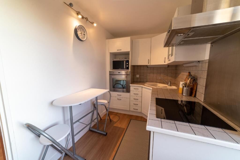A compact kitchen with white cabinets, a microwave, oven, and a stainless steel range hood. There is a small bar stool and a clock on the wall.