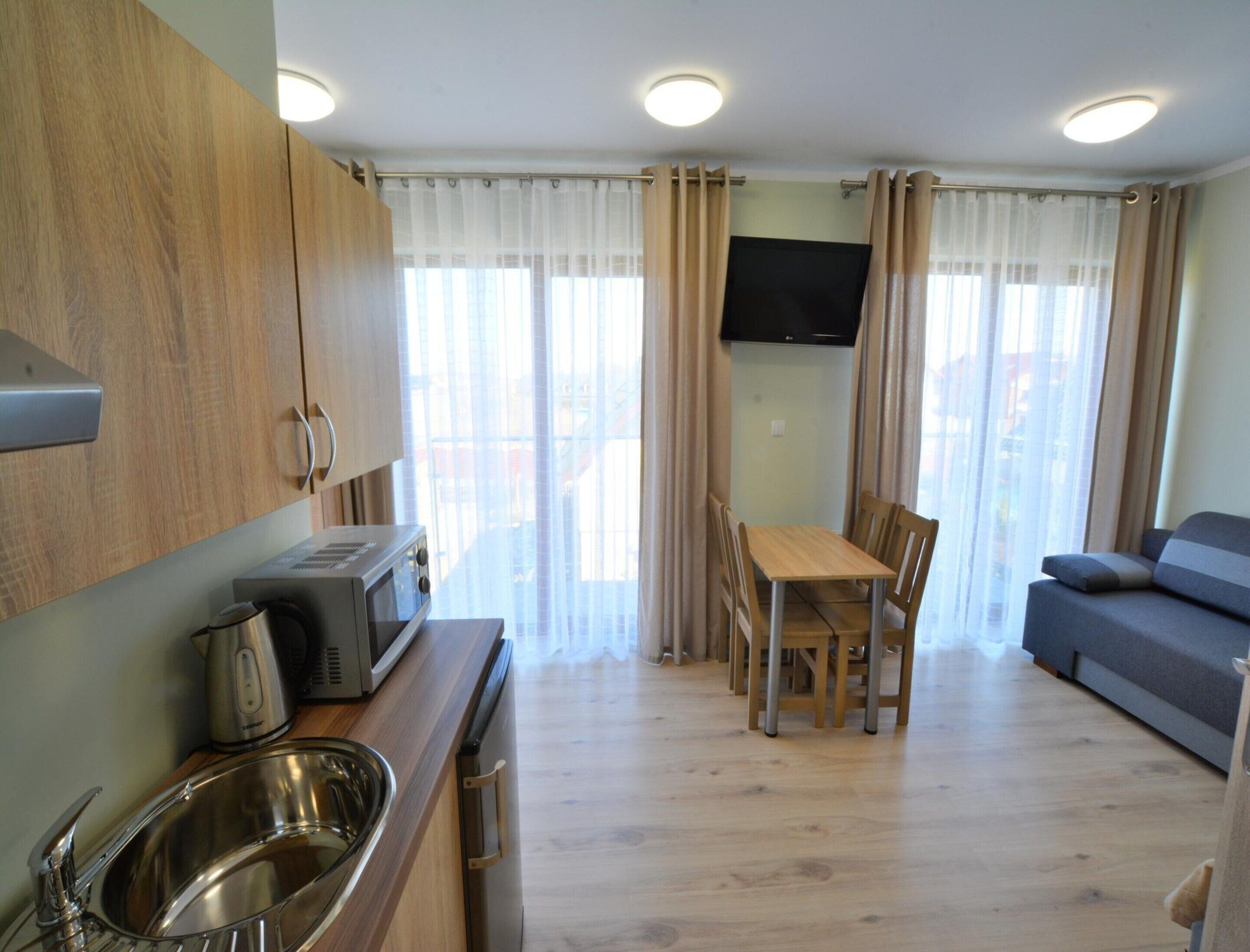 A compact kitchen with a microwave, kettle, and sink, a dining area with chairs and a table, and a sofa in a bright room with curtains and a TV.
