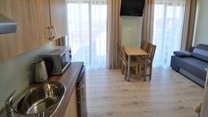 Eine kleine Küche mit Mikrowelle, Wasserkocher und Spüle, ein Essbereich mit Stühlen und Tisch, und ein Sofa in einem hellen Raum mit Vorhängen und einem Fernseher.