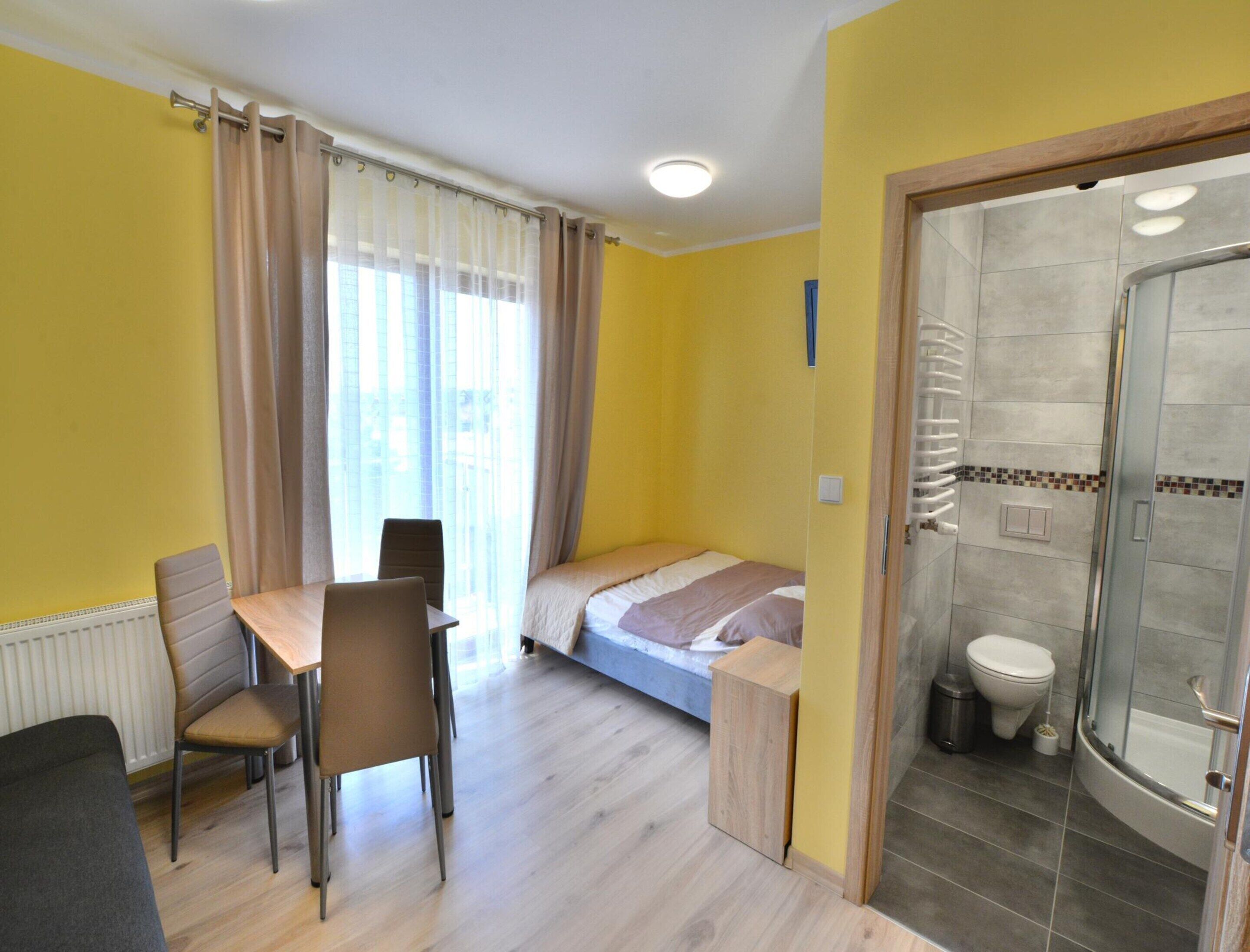 Une chambre d’hôtel de taille réduite, comprenant un lit, une petite table avec deux chaises, un espace douche et une fenêtre avec des rideaux.