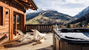 A wooden cabin with a hot tub and deck chairs overlooking a mountainous landscape.