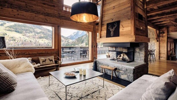 A cozy cabin interior with a stone fireplace, wooden walls, a sofa, and a dining area with a view of mountains.