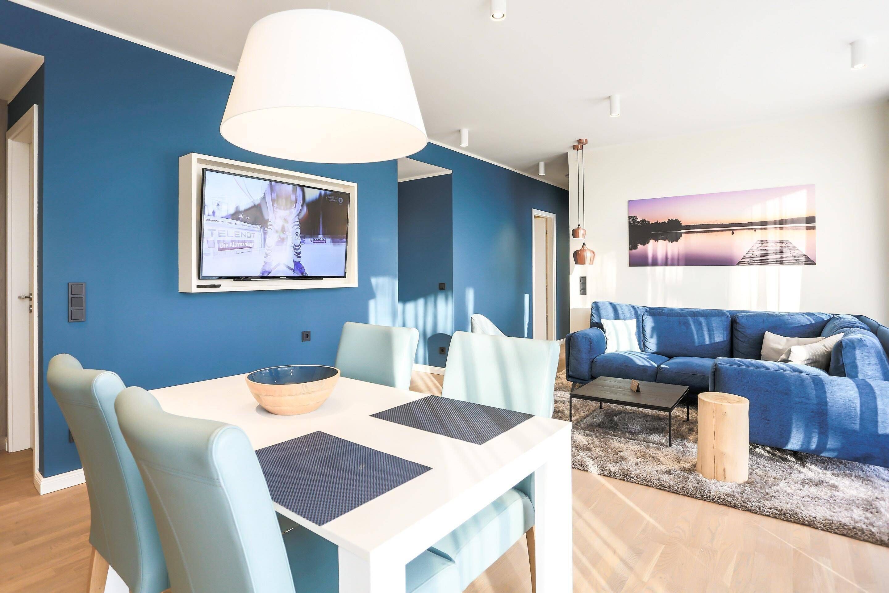 A modern living room with a blue sofa, a dining table with chairs, and a TV mounted on the wall.