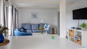 Ett modernt vardagsrum med en vit soffa, randiga kuddar, ett vitt soffbord, en hängande lampa och en väggmonterad TV.