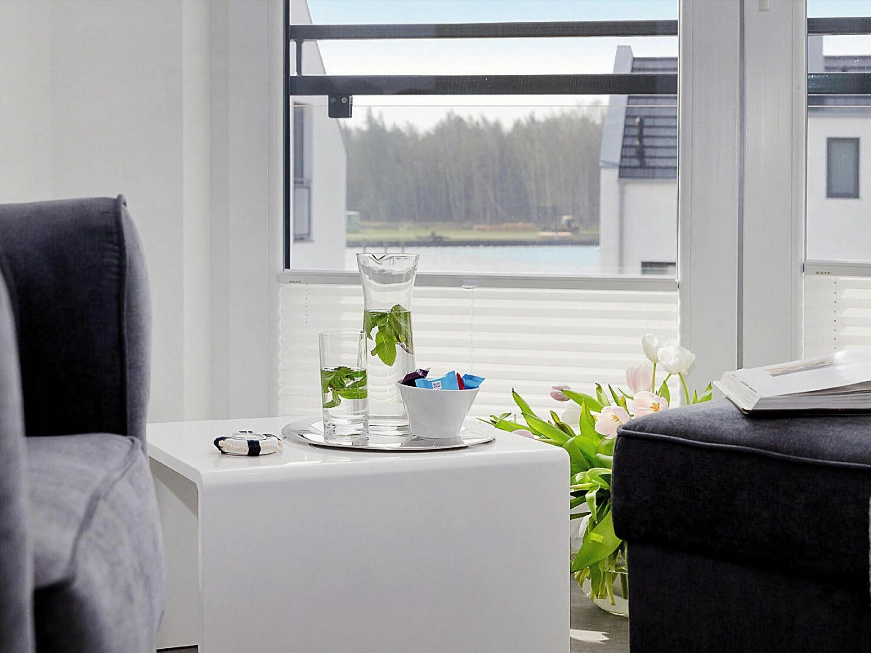 Een moderne kamer met een groot raam, een witte tafel met een bloevas, een glas water en een schaaltje met snacks.