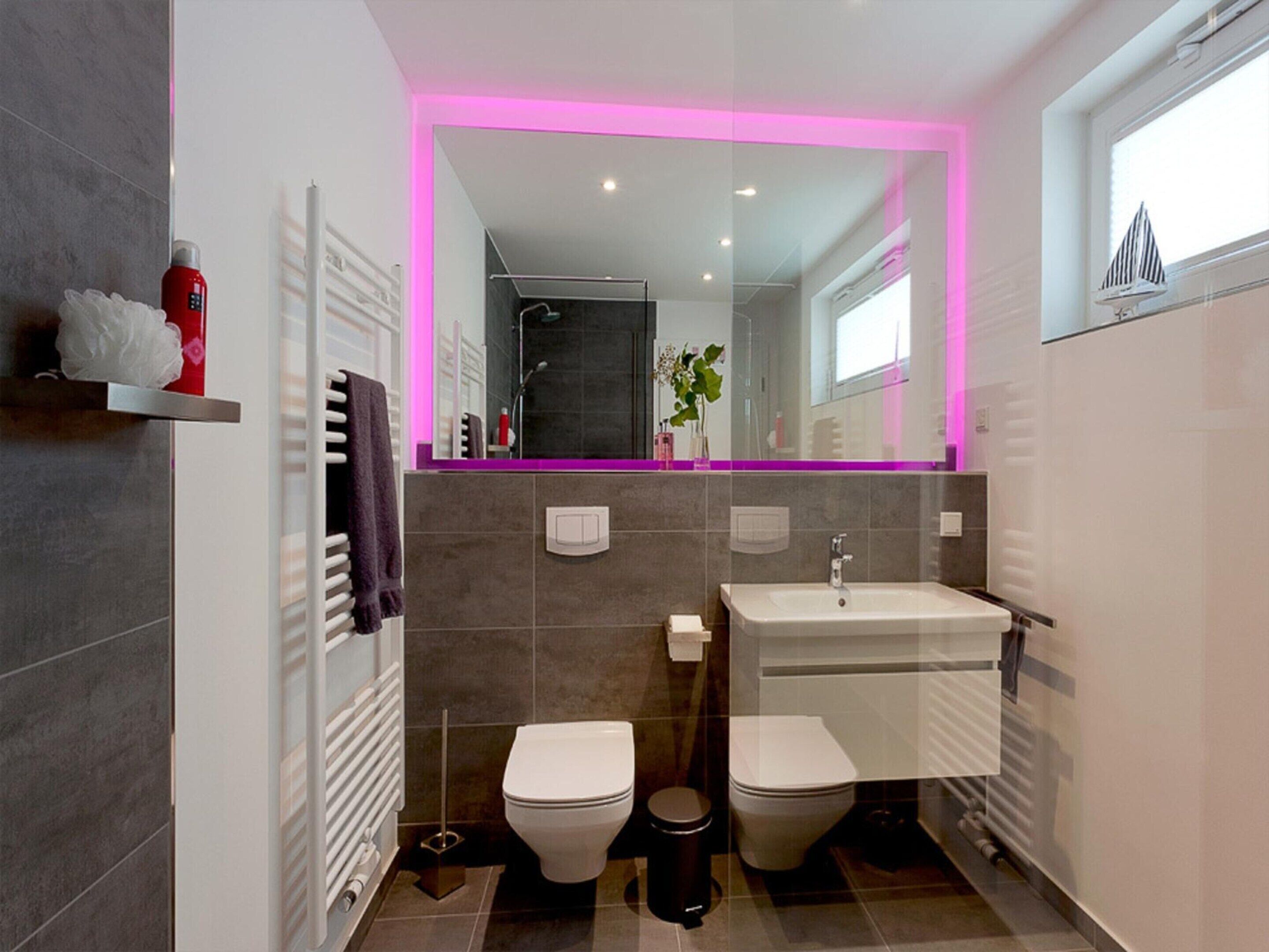 A modern bathroom with a dual toilet setup, a white sink, and a mirror with pink lighting.