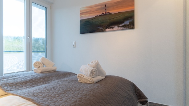 A hotel room with a bed, a painting of a lighthouse, and a balcony with a view of greenery. - Wohnung in St. Peter Ording (St. Peter-Ording)