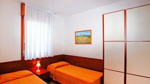 Una camera da letto con due letti singoli, una testiera in legno, un tavolino con una lampada, una finestra con tende bianche e un quadro appeso al muro.