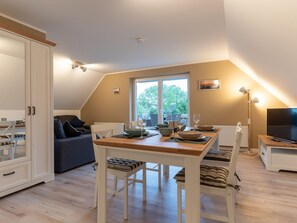 A cozy, well-lit room with a dining table set for four, a sofa, a TV, and a large window offering a view of the outdoors. - Wohnung in St.peter Ording (St. Peter-Ording)