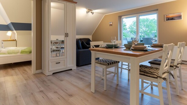 A room with a dining table set for four, a sofa, a wardrobe, and a bed with a nightstand. - Wohnung in St.peter Ording (St. Peter-Ording)