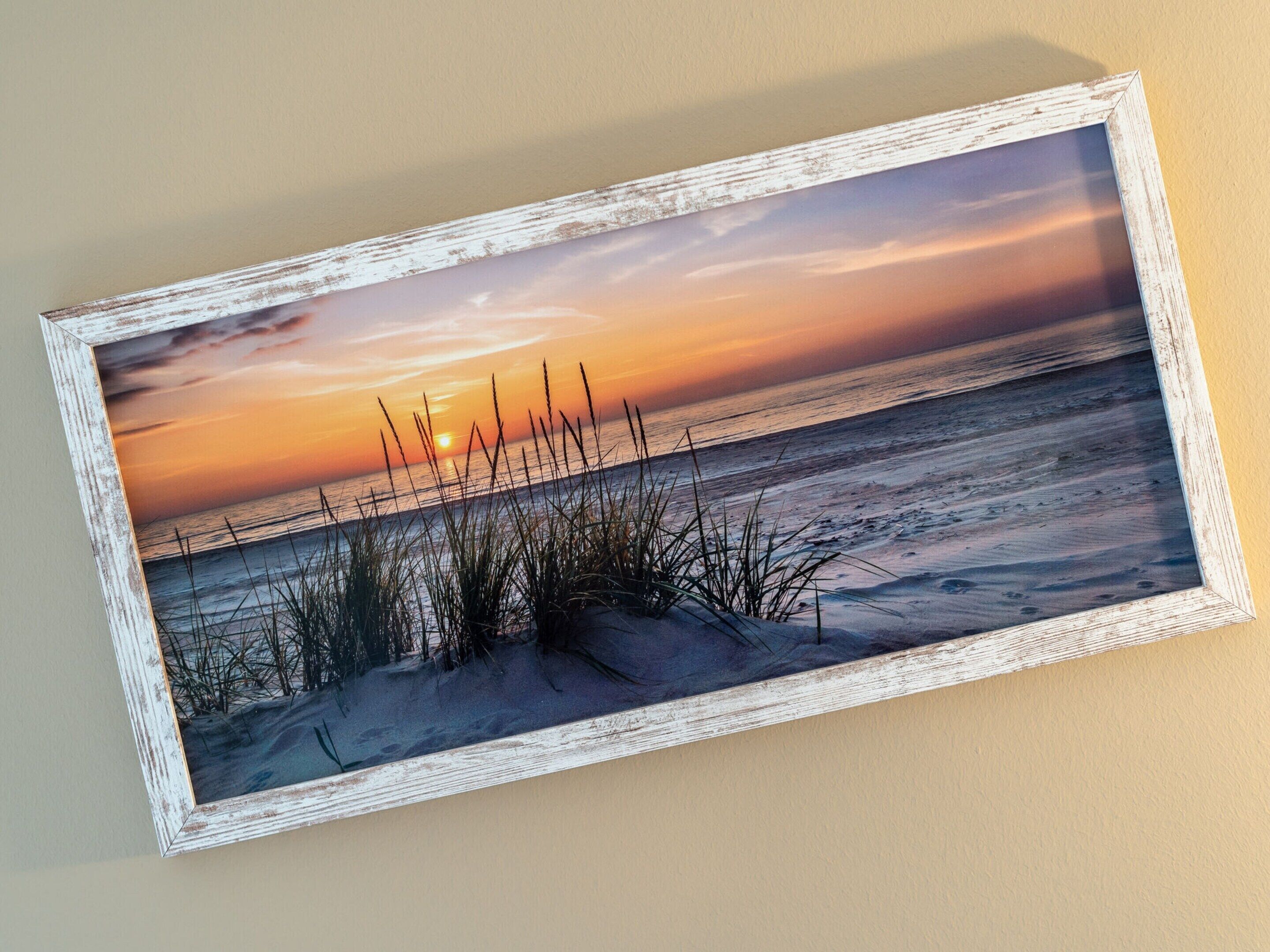 A framed landscape painting of a sunset over the ocean with grass in the foreground.