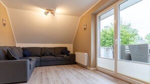 A modern living room with a sectional sofa, a radiator, and a large sliding glass door leading to an outdoor area. - Wohnung in St.peter Ording (St. Peter-Ording)