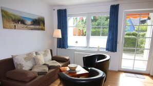 Ein Wohnzimmer mit einem braunen Sofa, zwei schwarzen Sesseln, einem Tisch mit einer Schale, einer Lampe und einem Fenster mit Vorhängen.
